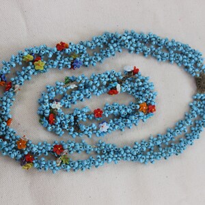 May include: A blue beaded necklace and bracelet set with colorful flower accents. The necklace is made of multiple strands of small blue beads and the bracelet is a single strand.