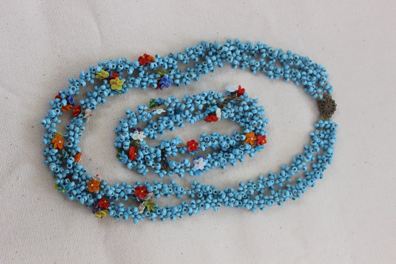 May include: A blue beaded necklace and bracelet set with colorful flower accents. The necklace is made of multiple strands of small blue beads and the bracelet is a single strand.