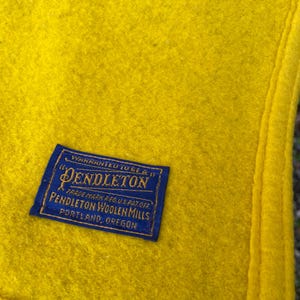 May include: Close-up of a bright yellow wool blanket with a navy blue label. The label reads "Pendleton Woolen Mills, Portland, Oregon". The blanket has a stitched edge and a soft, textured appearance.