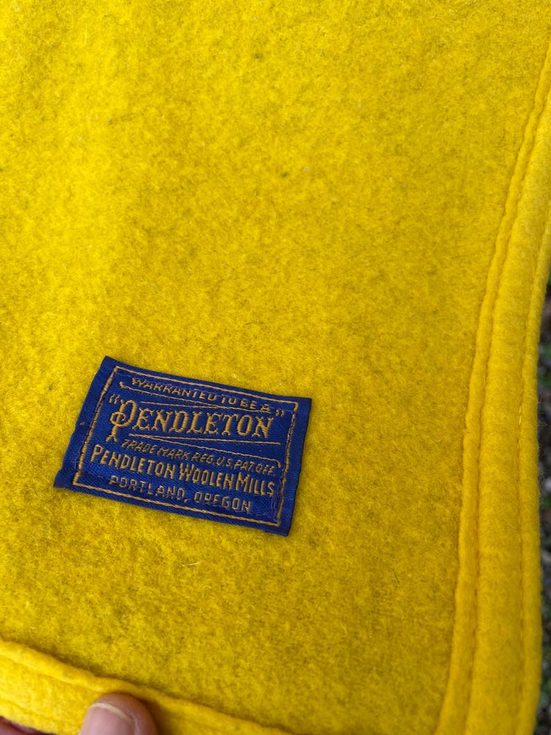 May include: Close-up of a bright yellow wool blanket with a navy blue label. The label reads "Pendleton Woolen Mills, Portland, Oregon". The blanket has a stitched edge and a soft, textured appearance.