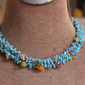 May include: A blue beaded necklace with colorful flower accents. The necklace is made of small, round beads and features a variety of colors, including red, orange, yellow, and white.