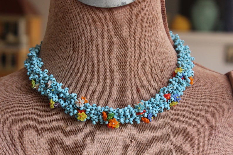 May include: A blue beaded necklace with colorful flower accents. The necklace is made of small, round beads and features a variety of colors, including red, orange, yellow, and white.