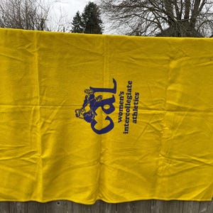 May include: A bright yellow blanket with the text "Cal women's intercollegiate athletics" in navy blue. The blanket is rectangular and appears to be made of a soft material. The blanket is hanging on a wooden fence.