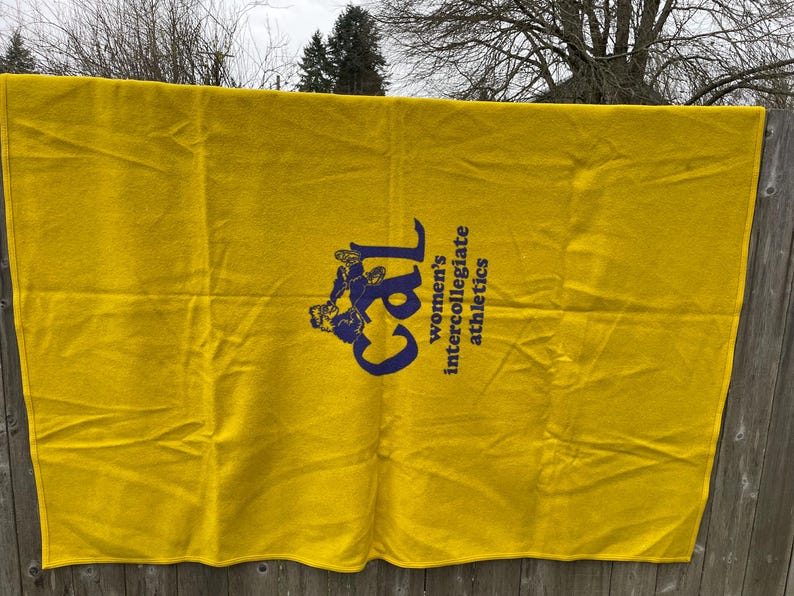 May include: A bright yellow blanket with the text "Cal women's intercollegiate athletics" in navy blue. The blanket is rectangular and appears to be made of a soft material. The blanket is hanging on a wooden fence.