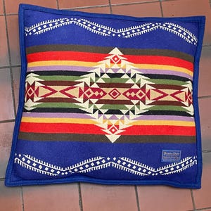 May include: A square, blue pillow with a colorful geometric pattern. The pillow has a blue border and a central design featuring red, yellow, green, and brown stripes. The pillow has a tag that says "Beaver State".