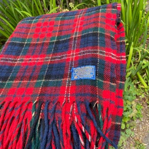 May include: A folded wool blanket featuring a red, dark blue, and dark green plaid pattern with a fringed edge. A blue rectangular label is stitched onto the fabric, displaying the text Pendleton, 100% Virgin Wool, and Made in U.S.A.