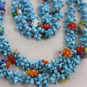 May include: A multi-strand necklace made of turquoise blue beads with colorful flower accents. The necklace is made of small, round beads and features a variety of colors, including red, orange, yellow, green, blue, and purple.