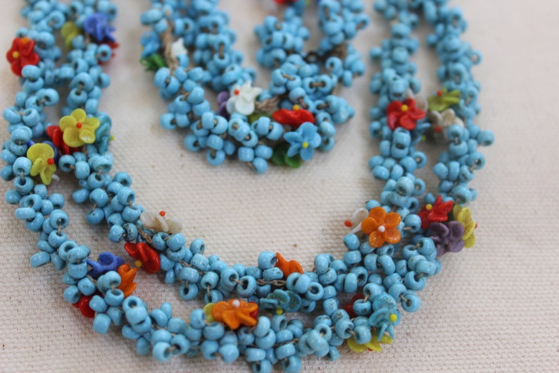 May include: A multi-strand necklace made of turquoise blue beads with colorful flower accents. The necklace is made of small, round beads and features a variety of colors, including red, orange, yellow, green, blue, and purple.