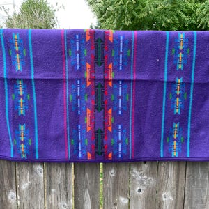 May include: A rectangular purple blanket featuring a central vertical pattern of geometric shapes in orange, green, and blue. The design includes symmetrical columns of repeating arrow-like motifs and thin vertical stripes in teal and magenta.
