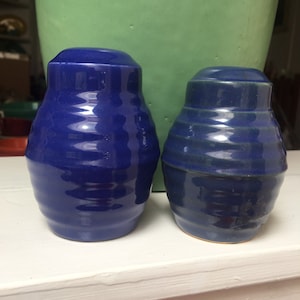 May include: Two ceramic salt and pepper shakers. The shaker on the left is a deep blue color. The shaker on the right is a darker blue with a green glaze.