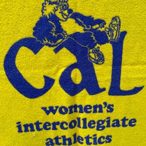 May include: Yellow fabric with a blue graphic of a cartoon figure and the word "Cal". Below, the text reads "women's intercollegiate athletics". The figure is wearing sneakers and is sitting on the letter "l".