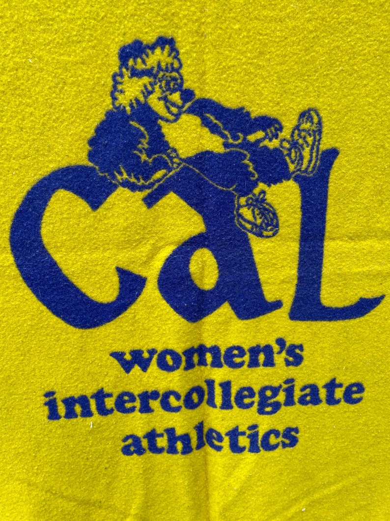 May include: Yellow fabric with a blue graphic of a cartoon figure and the word "Cal". Below, the text reads "women's intercollegiate athletics". The figure is wearing sneakers and is sitting on the letter "l".