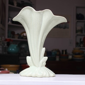 May include: A light green ceramic vase shaped like a lily flower. The vase has a smooth, textured surface and a wide opening at the top.