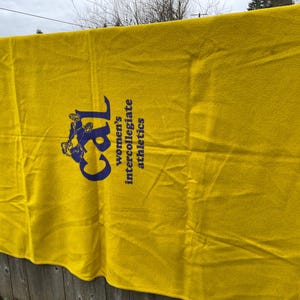 May include: A bright yellow blanket with the text "Cal women's intercollegiate athletics" in navy blue. The blanket is hanging on a wooden fence.