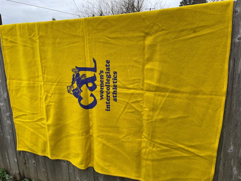 May include: A bright yellow blanket with the text "Cal women's intercollegiate athletics" in navy blue. The blanket is hanging on a wooden fence.