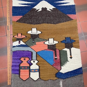 May include: A colorful woven tapestry featuring a mountain scene and figures in hats. The tapestry has a brown, blue, white, and purple color scheme. The tapestry is approximately 30 inches long.