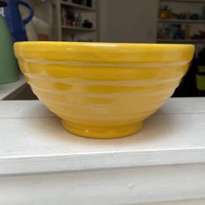 May include: A bright yellow ceramic bowl with a ribbed design. The bowl has a rounded shape and sits on a small base. The bowl is likely used for serving food or as a decorative piece.