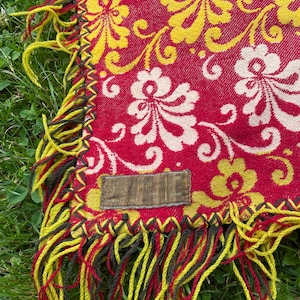 May include: A woven textile featuring a vibrant red background with a repeating yellow and cream floral damask pattern. The edges are finished with a dark decorative blanket stitch and long, multi-colored yellow and dark fringe tassels.