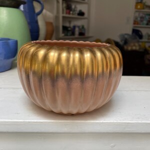 May include: A decorative, ribbed bowl with a gold-colored top and a pinkish-brown base. The bowl has a rounded shape and sits on a white surface. Other ceramic vases are visible in the background.