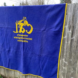 May include: A blue blanket with yellow trim and lettering. The blanket features the text "CSL" with a graphic of a person, and the words "women's intercollegiate athletics" in yellow.