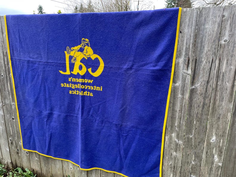 May include: A blue blanket with yellow trim and lettering. The blanket features the text "CSL" with a graphic of a person, and the words "women's intercollegiate athletics" in yellow.