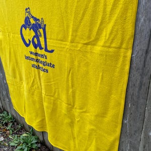 May include: A yellow blanket with the text "Cal women's intercollegiate athletics" in blue. The blanket is hanging on a wooden fence. The blanket has a rectangular shape.