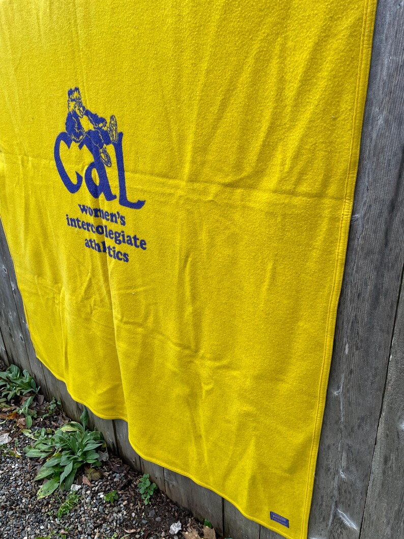 May include: A yellow blanket with the text "Cal women's intercollegiate athletics" in blue. The blanket is hanging on a wooden fence. The blanket has a rectangular shape.