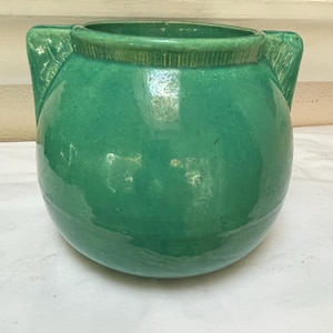 May include: A round, green ceramic pot with two side handles. The pot has a wide opening and a glossy finish. The color is a vibrant, solid green. The pot is likely for decorative or functional use.