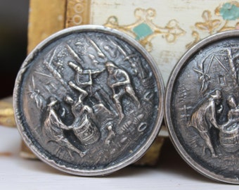 May include: Two silver-toned metal buttons with raised, detailed images of people working around a barrel. The buttons have a circular shape and a slightly raised rim.