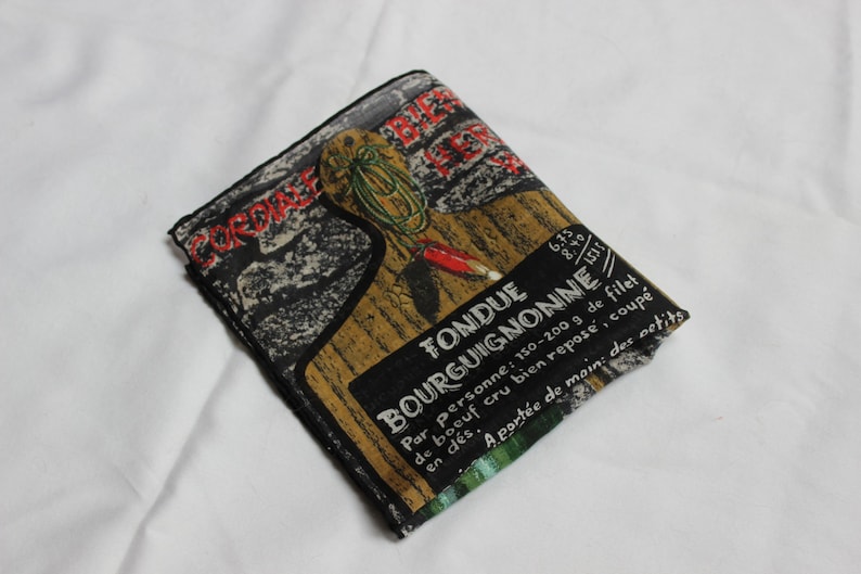May include: A black and brown fabric pouch with a vintage design featuring a fondue recipe. The text reads "Fondue Bourguignonne" and includes instructions for preparing the dish. The pouch is folded and has a green and white striped border.