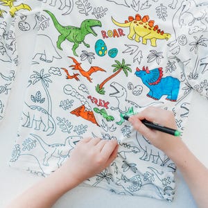 May include: A white long-sleeved shirt with a dinosaur theme. The shirt is designed for coloring with markers or crayons. The shirt features a variety of dinosaurs, including a T-Rex, a Triceratops, and a Stegosaurus. The shirt also includes the words "ROAR" and "TREX".
