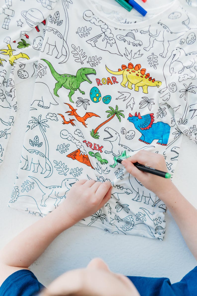 May include: A white long-sleeved shirt with a dinosaur theme. The shirt is designed for coloring with markers or crayons. The shirt features a variety of dinosaurs, including a T-Rex, a Triceratops, and a Stegosaurus. The shirt also includes the words "ROAR" and "TREX".