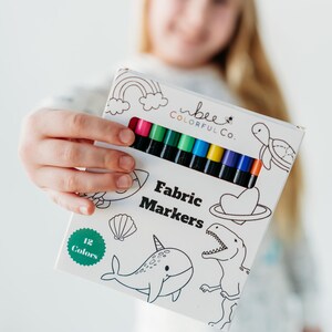 Young girl holding a box of Bee Colorful Co. fabric markers, featuring 12 rainbow colors. She smiles, showcasing the set designed for coloring pajamas and fabric crafts. These non-toxic markers, perfect for kids, are ideal for personalizing clothing, t-shirts, and canvas bags. The fine line tips allow for detailed designs, encouraging creative play and DIY projects. This image highlights the joy of using fabric markers for personalized gifts and artistic expression.