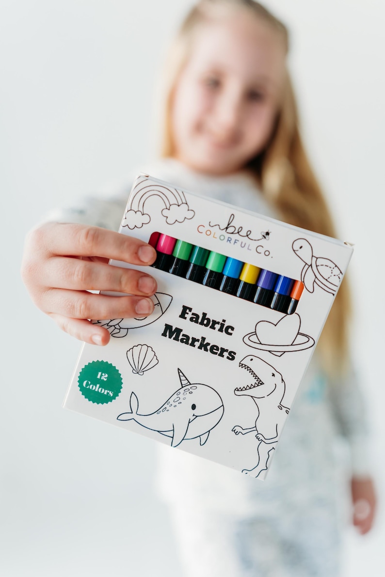 Young girl holding a box of Bee Colorful Co. fabric markers, featuring 12 rainbow colors. She smiles, showcasing the set designed for coloring pajamas and fabric crafts. These non-toxic markers, perfect for kids, are ideal for personalizing clothing, t-shirts, and canvas bags. The fine line tips allow for detailed designs, encouraging creative play and DIY projects. This image highlights the joy of using fabric markers for personalized gifts and artistic expression.