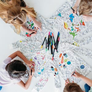 May include: Four children are coloring on a white fabric with colorful markers. The fabric has a variety of designs, including dinosaurs, space, and other animals. The fabric is spread out on a white surface.