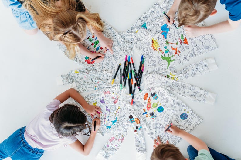 May include: Four children are coloring on a white fabric with colorful markers. The fabric has a variety of designs, including dinosaurs, space, and other animals. The fabric is spread out on a white surface.