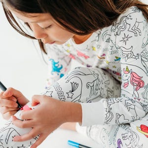 Puede incluir: Una persona joven lleva una pijama blanca con un dise&ntilde;o de unicornio y estrellas y est&aacute; coloreando la tela con un marcador negro. La pijama est&aacute; dise&ntilde;ada para ser coloreada.