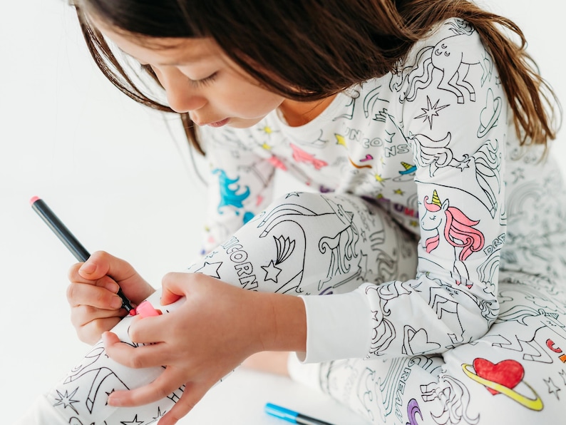 Puede incluir: Una persona joven lleva una pijama blanca con un dise&ntilde;o de unicornio y estrellas y est&aacute; coloreando la tela con un marcador negro. La pijama est&aacute; dise&ntilde;ada para ser coloreada.