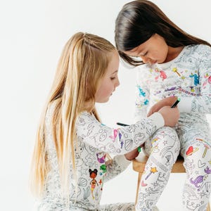 Puede incluir: Dos ni&ntilde;os con pijamas blancos con dibujos coloridos de sirenas, unicornios y otras criaturas. Los pijamas est&aacute;n dise&ntilde;ados para ser coloreados.