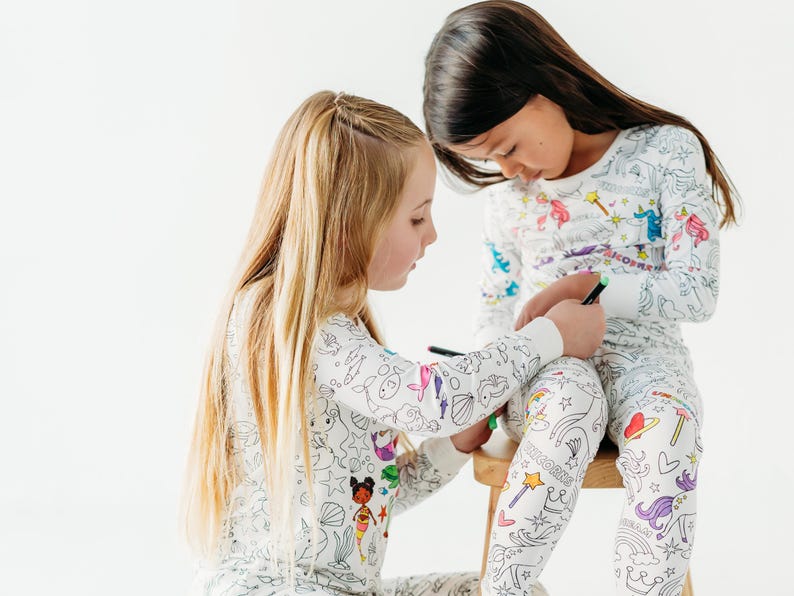 Puede incluir: Dos ni&ntilde;os con pijamas blancos con dibujos coloridos de sirenas, unicornios y otras criaturas. Los pijamas est&aacute;n dise&ntilde;ados para ser coloreados.