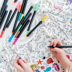 May include: A white t-shirt with a mermaid and sea creature design that is ready to be colored in with markers. The t-shirt is lying flat on a white surface with a variety of colored markers scattered around it.