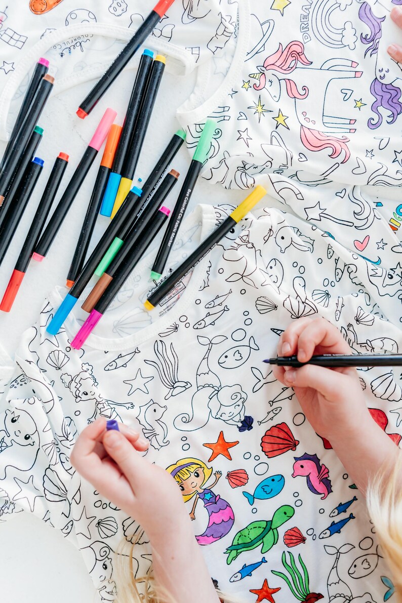 May include: A white t-shirt with a mermaid and sea creature design that is ready to be colored in with markers. The t-shirt is lying flat on a white surface with a variety of colored markers scattered around it.