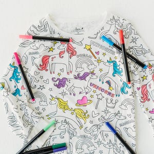 May include: White long-sleeved shirt with a unicorn and rainbow design. The shirt is ready to be colored in with the included markers. The markers are black, blue, green, pink, and red.