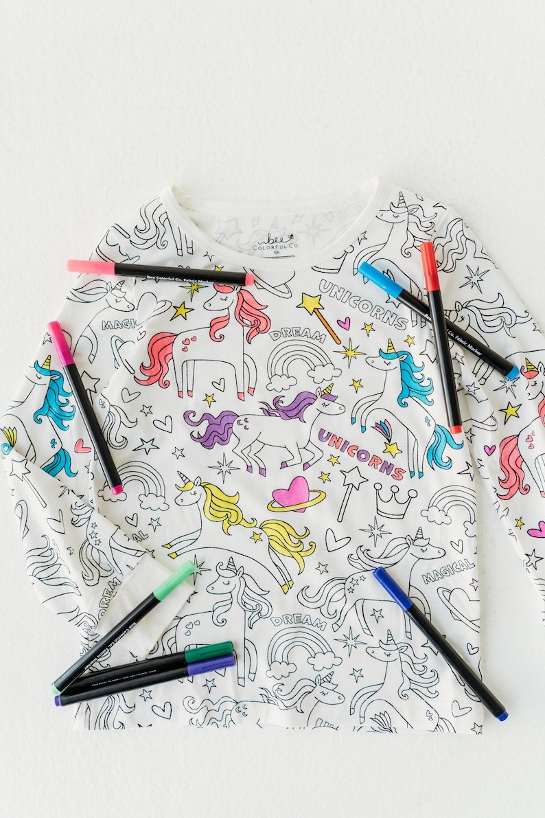 May include: White long-sleeved shirt with a unicorn and rainbow design. The shirt is ready to be colored in with the included markers. The markers are black, blue, green, pink, and red.