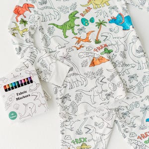 May include: White pajamas with a dinosaur theme. The pajamas have outlines of dinosaurs, plants, and eggs that can be colored in. A box of 12 fabric markers is included. The box has a blue and white design with a narwhal, a rocket, and a seashell.