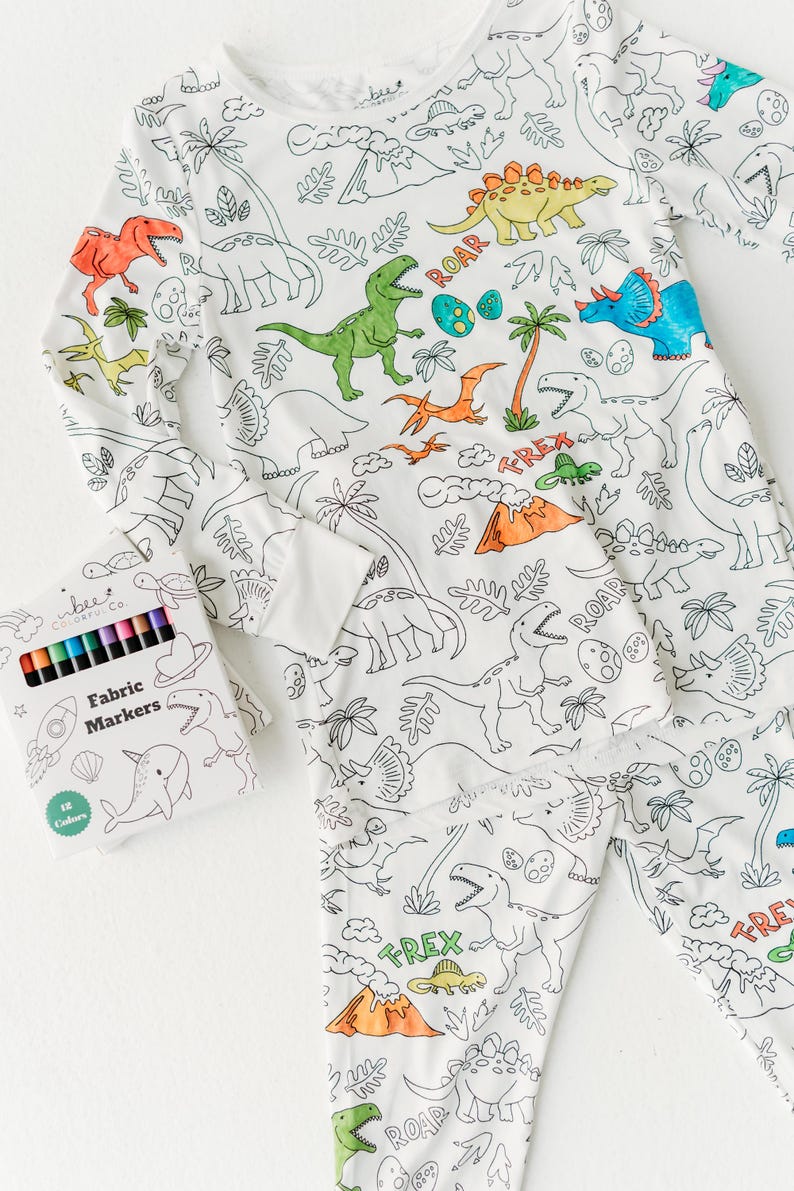 May include: White pajamas with a dinosaur theme. The pajamas have outlines of dinosaurs, plants, and eggs that can be colored in. A box of 12 fabric markers is included. The box has a blue and white design with a narwhal, a rocket, and a seashell.