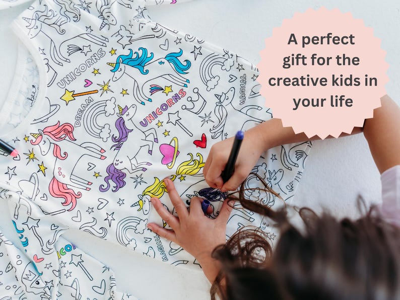 Image shows a young girl joyfully coloring her unicorn pajamas with vibrant fabric markers. The photo includes the text 'A perfect gift for the creative kids in your life,' highlighting the imaginative play and personalized gift aspect of these pajamas.