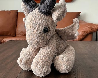 May include: A plush dragon toy crafted from soft, textured beige yarn. The creature features two prominent dark gray horns on its head, large dark eyes, pointed ears, and small wings on its back. It is depicted in a seated position with visible paws.