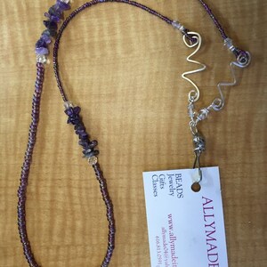 May include: A purple and silver beaded necklace with amethyst stones and a silver wire design. The necklace is hanging from a business card that reads "ALLYMADE Beads Jewelry Gifts Classes www.allymadeit.com allymade@yahoo.com 616.813.0591"