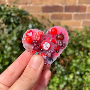 Peut inclure: Un objet en résine en forme de cœur avec des fruits miniatures incrustés, des coccinelles et d'autres éléments décoratifs. La résine est rose et translucide, avec des accents rouges et blancs. L'objet est tenu dans une main.